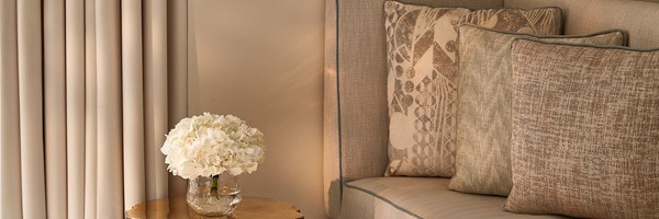 Close-up of a cushioned bench with textured beige pillows beside a side table holding white flowers.