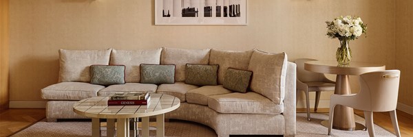 Neutral-toned lounge area featuring a curved upholstered sofa with blue cushions, a tiled coffee table with books, and a round dining table with white flowers in a glass vase.