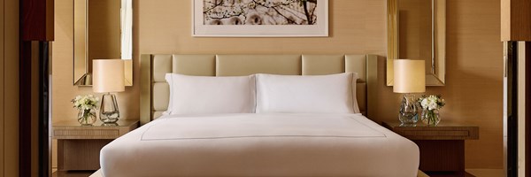 King-sized bed with a padded cream headboard, white linens, and two bedside tables topped with floral arrangements and glass lamps, beneath a framed cherry blossom print.
