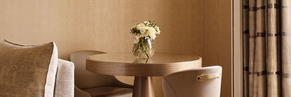 Small dining nook with a circular wood table, two beige leather chairs, and a clear vase of white flowers, beside patterned beige curtains.