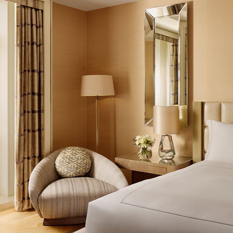 Cozy bedroom corner with a rounded striped armchair, geometric cushion, mirrored wall panel, and floor lamp beside a bedside table with flowers.