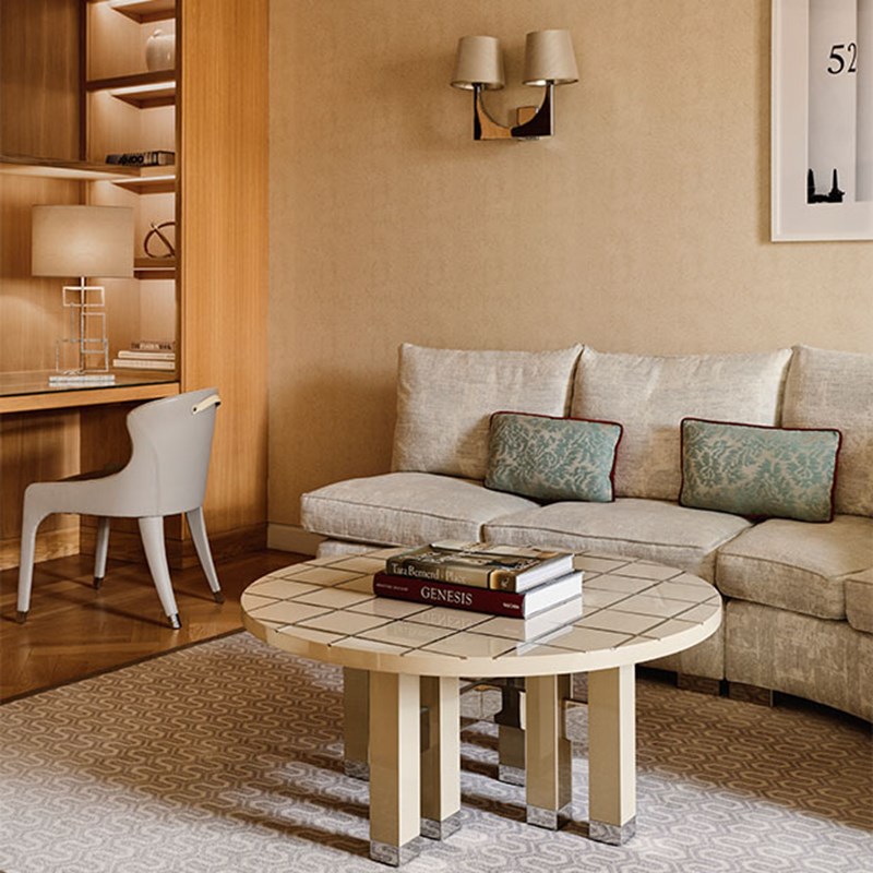 Neutral lounge with a curved sofa, pale blue cushions, tiled coffee table with art books, and a writing desk with shelves and a modern white chair.