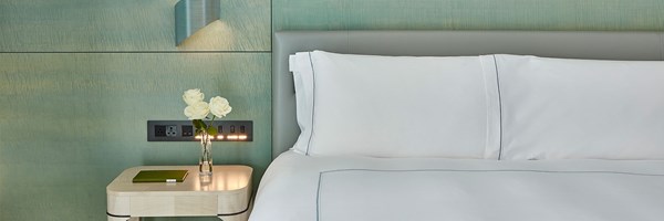 Double bed made up with white bed sheets, vase of three white roses on a bedside table, green coloured wall behind