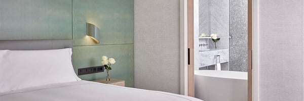 View of one side of a double bed made up with white bed sheets with grey lining,  bed side table with 3 white roses in a vase, green coloured wall, and view into marble bathroom with large bath