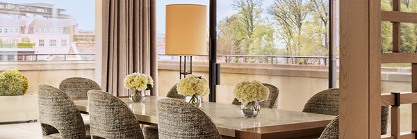 Bright dining room with a long marble table, soft upholstered chairs, and floor-to-ceiling glass doors opening to a view of trees and rooftops.