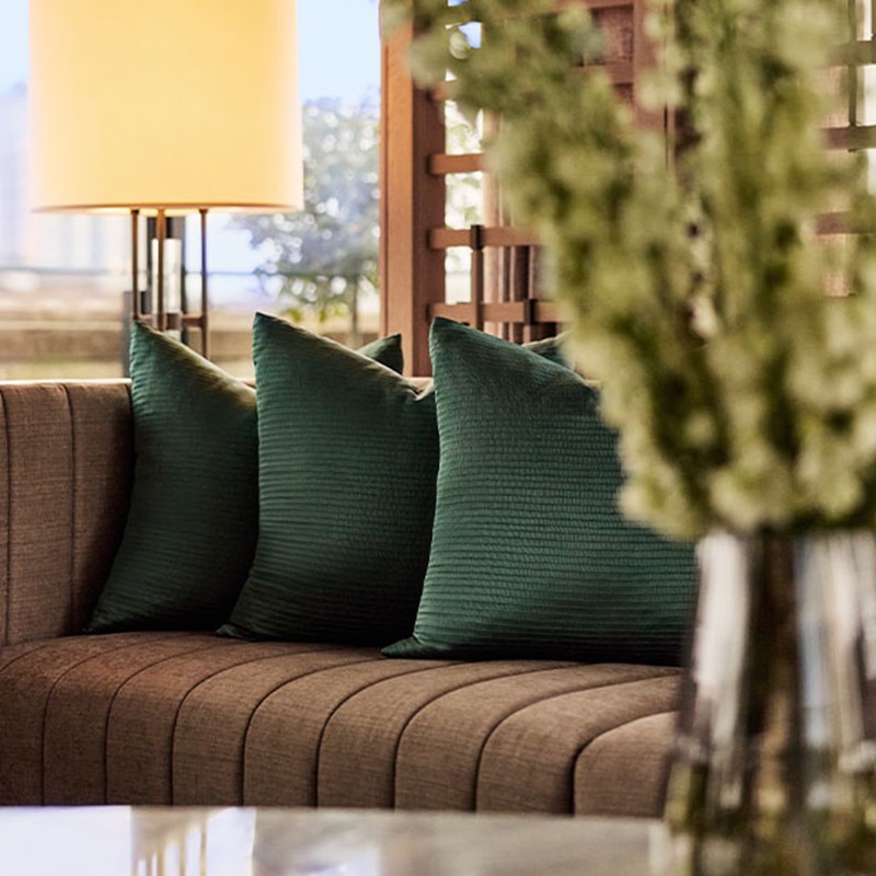 Close-up of plush green pillows on a curved sofa, with soft lighting and a glass vase of white flowers in the foreground.