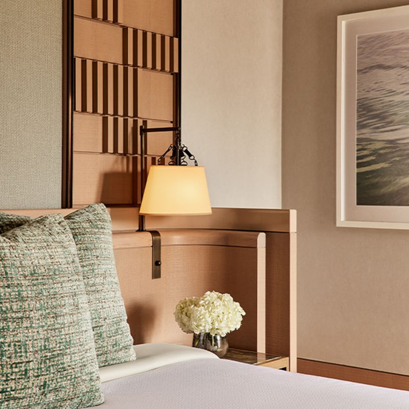Detail of a bedside area with a soft-glow wall lamp, green accent pillows, and a small vase of white hydrangeas on a nightstand.