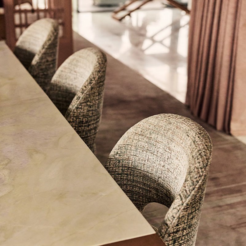 Detail view of curved upholstered dining chairs in textured fabric beside a pale marble dining table, with soft light and neutral tones.
