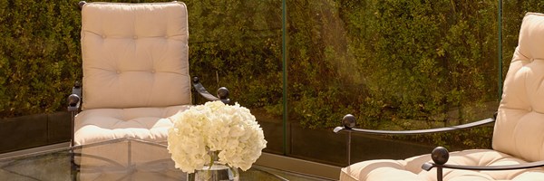 An intimate outdoor garden space with cushioned iron chairs and a glass-topped table, framed by manicured greenery and soft natural light.