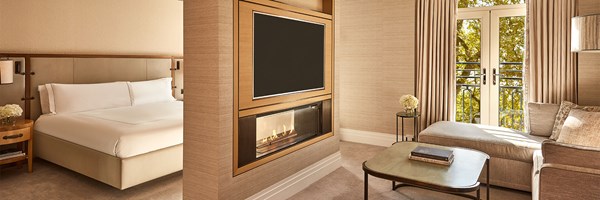 A spacious junior suite featuring a king-size bed with crisp white linens, a seating area with a cream L-shaped sofa, a fireplace beneath a flat-screen TV, and French doors opening to a balcony with leafy views.