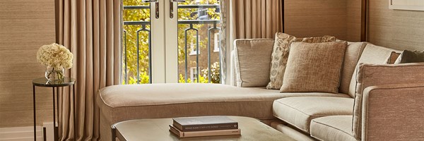 Detail of the suite’s seating area showing a soft beige corner sofa with decorative cushions, a side table with white hydrangeas, and French doors revealing a balcony with views of tree-lined streets.
