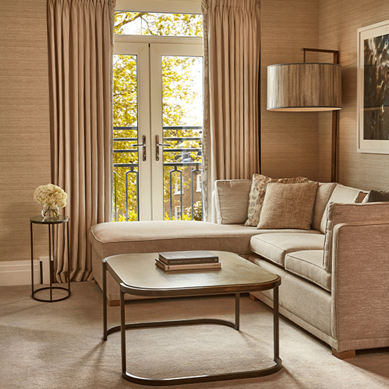 Cozy lounge corner of the Deluxe Junior Suite with a curved coffee table, neutral-toned sofa, side flowers, and full-length curtains framing a balcony door with tree views.