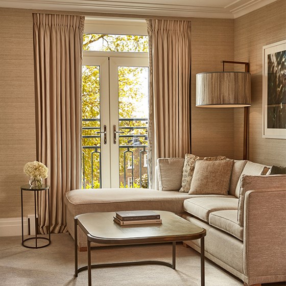 Elegant sitting area with L-shaped sofa, floor lamp, books on coffee table, and French doors opening to leafy city views.