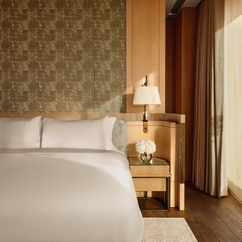 A serene bedroom with a luxurious white bed, a warm wood-paneled headboard, and a soft gold-accented wall lamp, bathed in golden light from sheer curtains beside floor-to-ceiling windows.