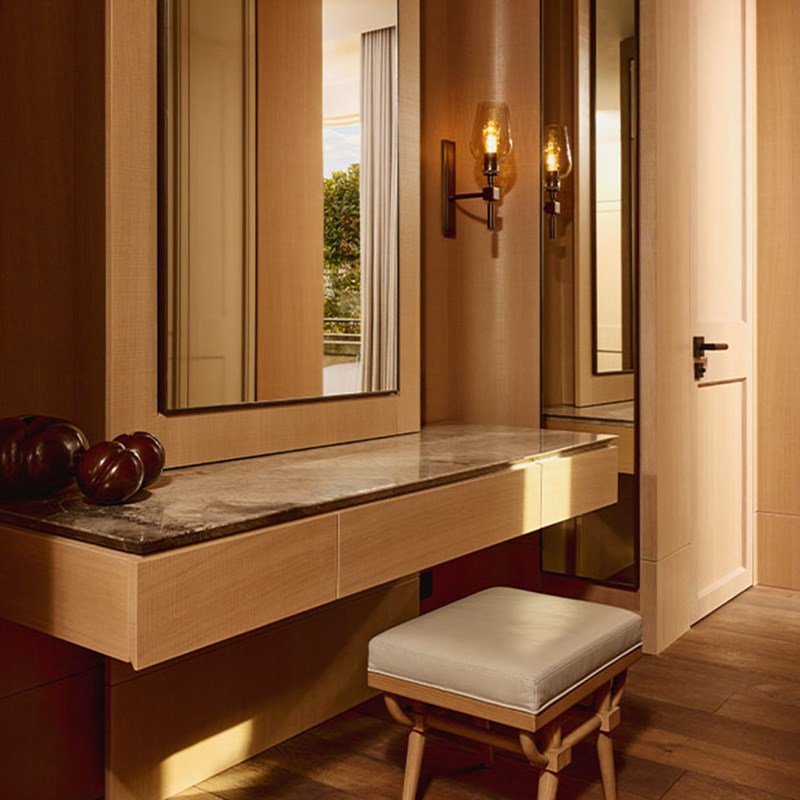 A refined dressing table with a floating marble countertop, large framed mirrors, and modern wall sconces, complemented by a simple cushioned stool and warm wooden flooring.