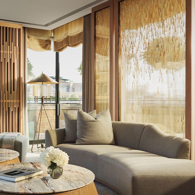 An elegant corner lounge area with a curved, soft-grey sofa, textured wooden screens, and shimmering gold-accented blinds, offering views to the terrace through large windows.