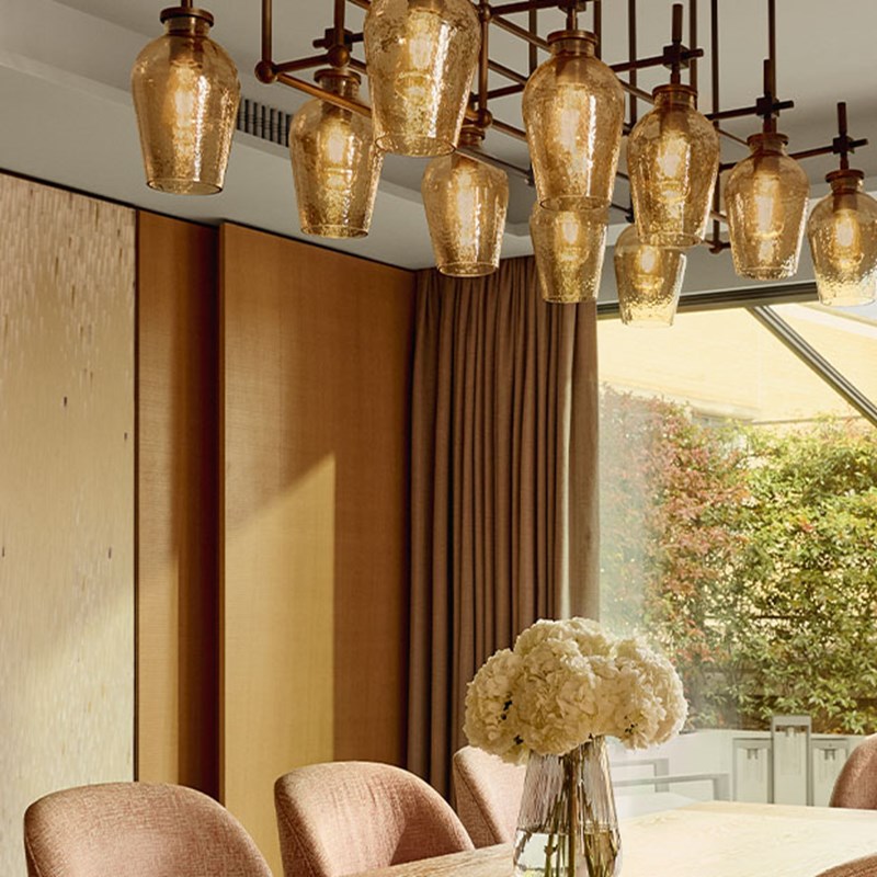 A sophisticated dining setting featuring a large cluster of amber-toned glass pendant lights suspended above a table adorned with a vase of white hydrangeas, framed by natural wood panels and floor-to-ceiling curtains.