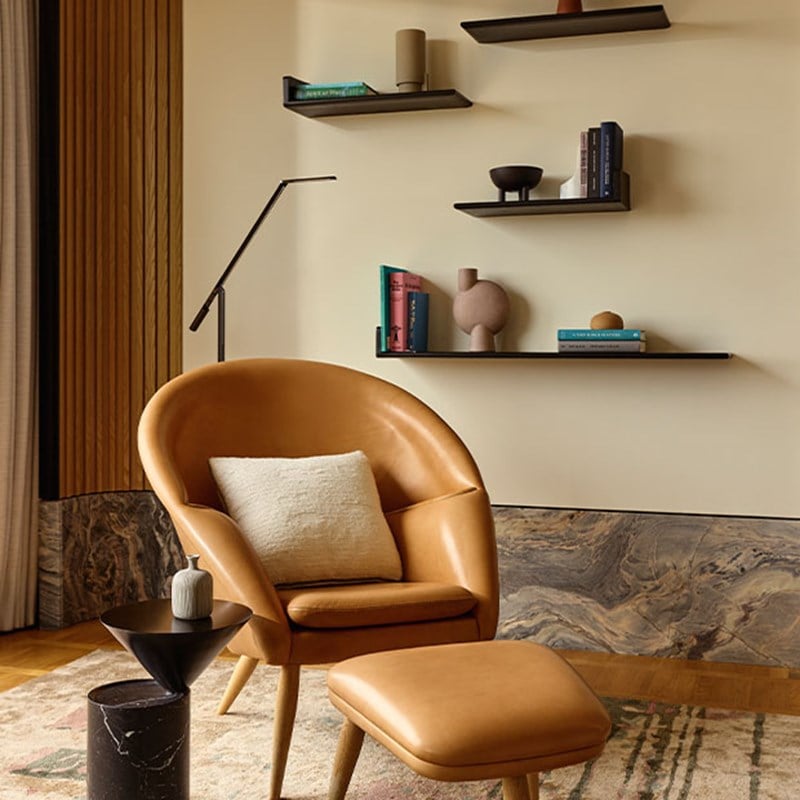Stylish reading nook with a caramel-toned leather chair and ottoman, black sculptural side tables, and floating shelves displaying books and modern decorative pieces against a cream wall.