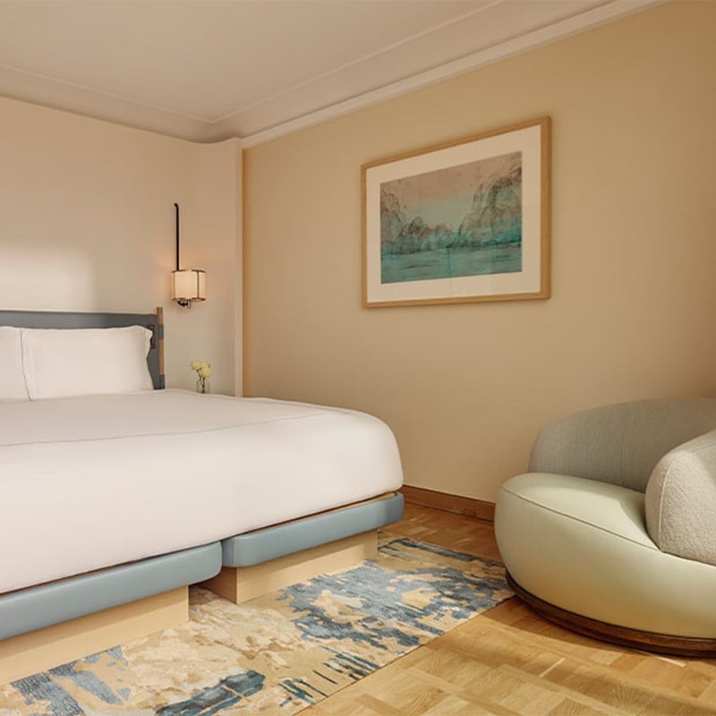 Calming bedroom with pale blue accents, a low-profile bed, modern sconce lighting, and a large framed seascape artwork above the headboard, complemented by a soft curved lounge chair.