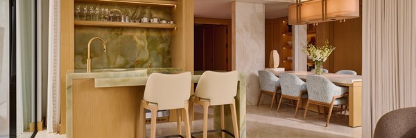 A refined open-plan bar and dining area, showcasing a striking green onyx bar counter, gold accents, and a marble-topped dining table surrounded by textured pastel chairs.
