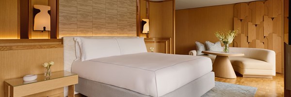 A tranquil bedroom with a crisp white king-size bed, soft lighting, and bespoke wood paneling, paired with a sculptural wall feature and a curved cream chaise.