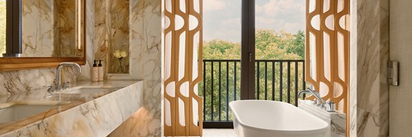 A luminous marble bathroom with a freestanding bathtub positioned before full-height windows, flanked by sculptural wooden screens and offering serene treetop views.