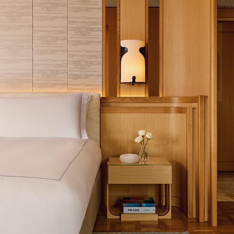 A serene bedroom corner with a neatly made bed in crisp white linens, a light oak bedside table adorned with minimalist décor, and a sculptural wall sconce softly illuminating the wood paneling.