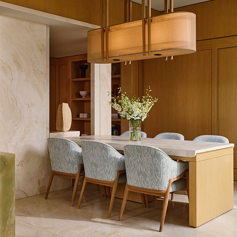 An elegant dining space featuring a light stone table surrounded by pale blue upholstered chairs, set against rich wood paneling and softened by a sculptural pendant light and fresh floral arrangement.