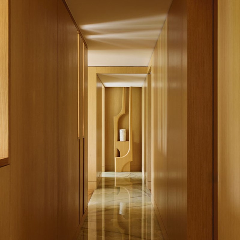 A minimalist corridor lined with warm wood panels and polished stone flooring, leading to an alcove with a sculptural shelf and a single decorative object, bathed in soft natural light.