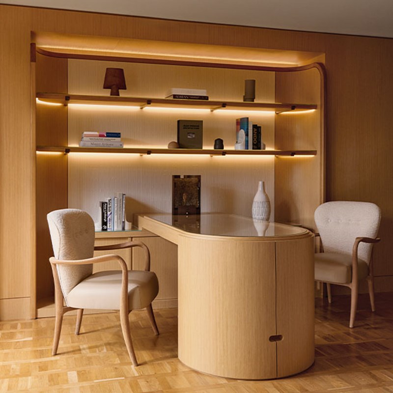 A refined workspace with a curved wooden desk, two cream armchairs, and softly lit open shelving displaying curated books and objets d’art, all in a palette of warm neutrals.