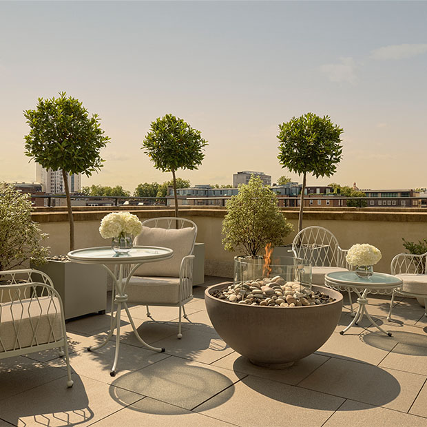 An expansive rooftop terrace with cream outdoor seating, manicured topiary trees, and a central fire pit surrounded by pebbles, offering views over the London skyline under a clear sky.