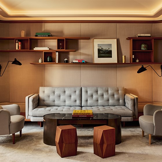 Stylish living room with a tufted grey sofa, armchairs, dark marble coffee table, geometric wood stools, and minimalist floating bookshelves on a warm-toned wall.