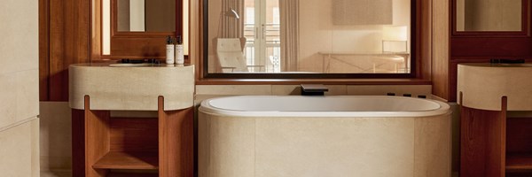 Elegant bathroom with a central bathtub, twin curved vanities in beige stone, and warm wood paneling with ambient lighting.