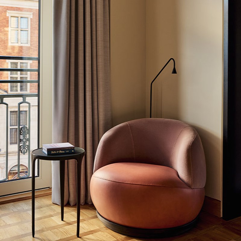 Close-up of a pink curved lounge chair beside a floor-to-ceiling window with sheer curtains and a side table with a book.