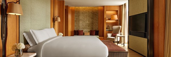 An expansive bedroom with contemporary lines, a luxurious upholstered bed, warm timber finishes, and a serene seating nook framed by natural textures and soft lighting.