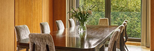 A sophisticated dining area with a polished dark-wood table, velvety taupe chairs, and floor-to-ceiling windows offering leafy views, styled with a centrepiece of fresh florals.