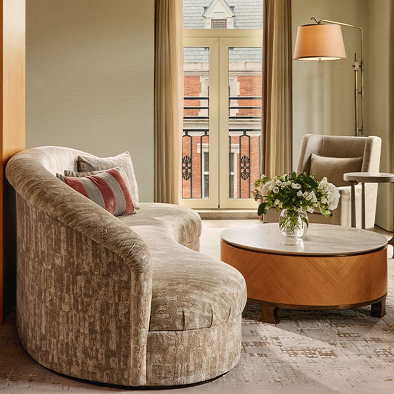 Refined seating area with a curved, upholstered sofa and round coffee table adorned with fresh flowers, set before tall windows with views of classic architecture.