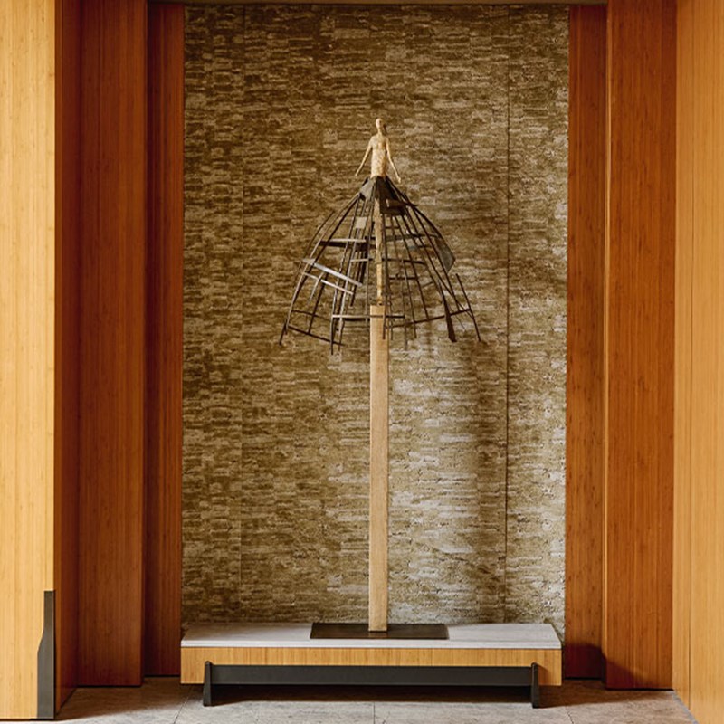 Sculpture of a slender human figure standing atop a tall pedestal, encased within a metal architectural frame resembling a skirt or dome, against a textured stone wall.