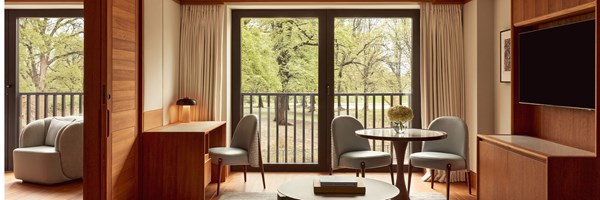 Bright suite interior with a small dining table, pale blue chairs, and a writing desk, overlooking a park through large glass doors.