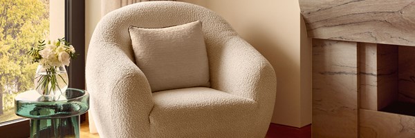 Close-up of a textured boucle armchair with a neutral cushion, beside a green glass side table and a vase of fresh white flowers.