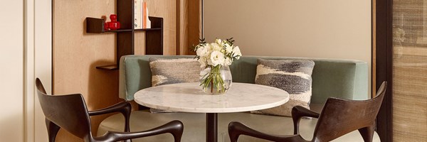 Intimate dining nook featuring a round marble table, curved mint green bench, dark wood chairs, and a vase of white flowers in a modern setting.