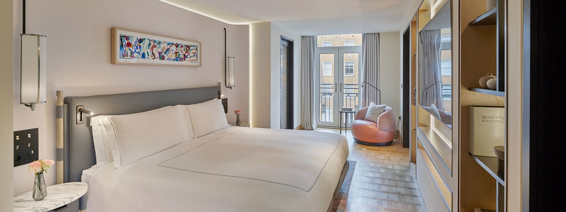 A view of a bedroom with a King Bed in the Knightsbridge Suite, with lots of daylight, and a pink armchair.