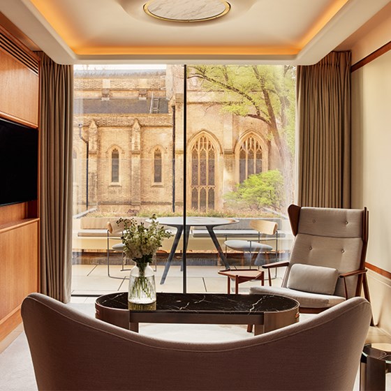Modern suite seating area with curved sofa, marble coffee table, and armchair facing floor-to-ceiling glass doors opening onto a terrace with a view of a historic stone church and leafy trees.