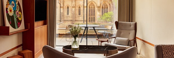Modern suite seating area with curved sofa, marble coffee table, and armchair facing floor-to-ceiling glass doors opening onto a terrace with a view of a historic stone church and leafy trees.