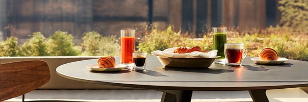 A close-up of a round outdoor table on the private terrace of the Terrace Junior Suite at The Berkeley, set with croissants, espresso, juice, and tea, with greenery and a blurred cityscape in the background