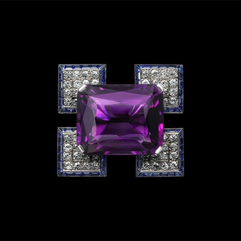 Cartier brooch with a large central purple amethyst, framed by four square clusters of diamonds bordered with sapphires, set against a black background.