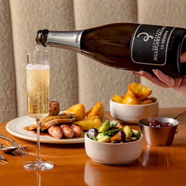 Sparkling wine being poured beside a festive roast with sausages, vegetables, and roast potatoes on a wooden table.
