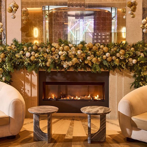 Festive hotel lounge with armchairs by a lit fireplace adorned with garlands and gold baubles.