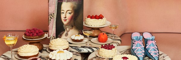 An array of ornate cakes topped with fruit sits on a patterned table beside a portrait of Marie Antoinette and a pair of pastel pink and blue ballet-style shoes.