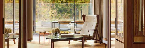 View through an elegant wooden-framed interior towards a private terrace, featuring a mid-century style armchair, low coffee table with fresh white hydrangeas, and floor-to-ceiling glass revealing lush autumn foliage beyond.